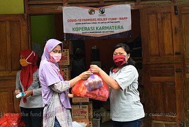 [Translate to fr:] Eine Frau mit Maske von der Kooperative Karmatera in Indonesien übergibt einer anderen Frau mit Maske zwei Tüten mit Grundnahrungsmitteln